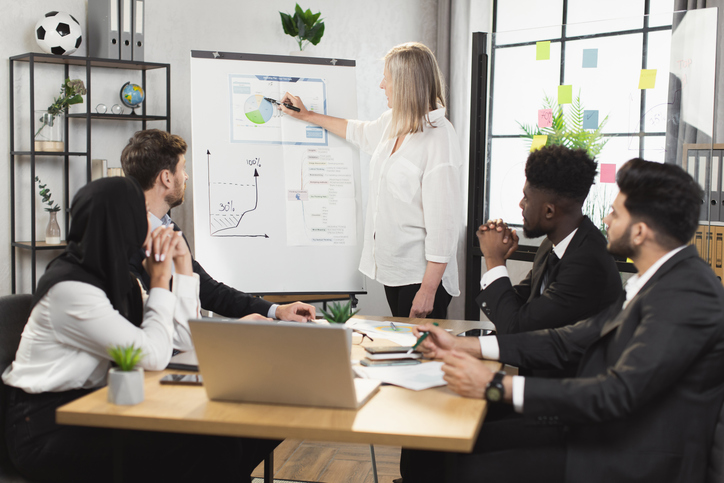 Caucasian grey haired woman presenting financial reports on flip chart to multicultural colleagues during conference at boardroom. Meeting of divers company workers.