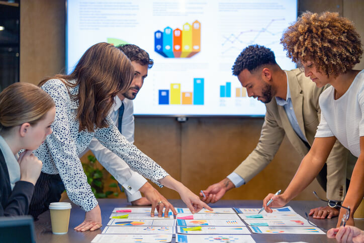 Paperwork and group of peoples hands on a board room table at a business presentation or seminar. The documents have financial or marketing figures, graphs and charts on them. People are pointing at the data. There is a T.V. with financial data in the background