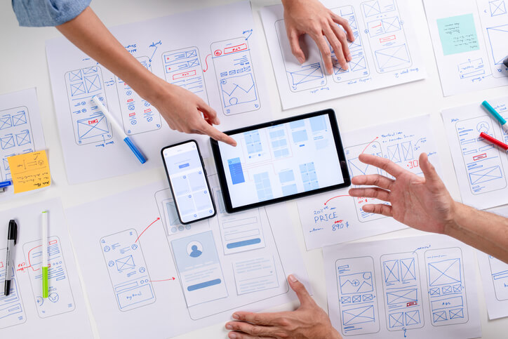 UX/UI designers discussing and brainstorming on wireframes for a website and mobile app prototype, surrounded by sketches of user flow and design tools, in the concept of website and mobile application design concept.